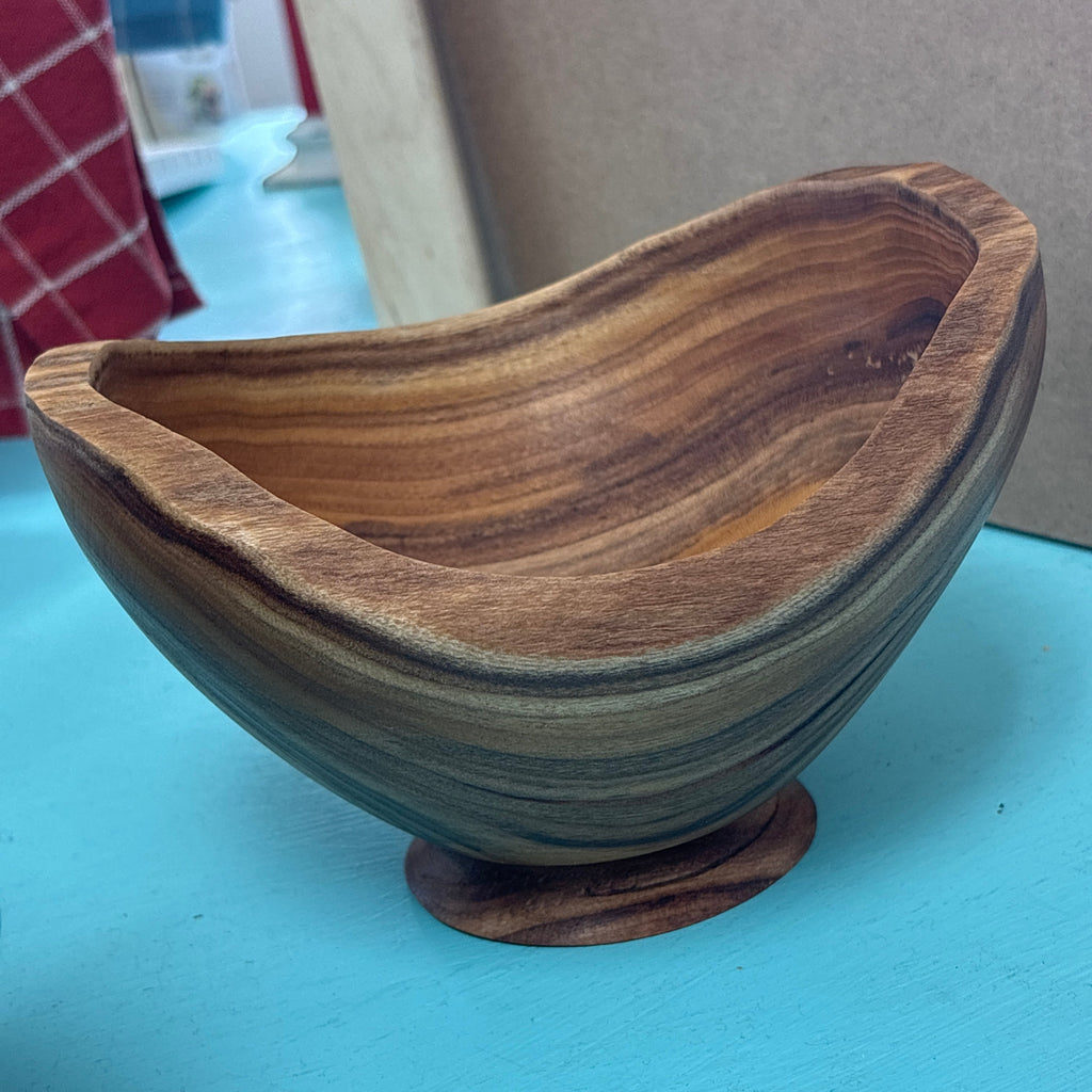 A handcrafted wooden bowl with a unique organic shape, featuring rich brown and tan wood grain patterns, resting on a light blue surface.