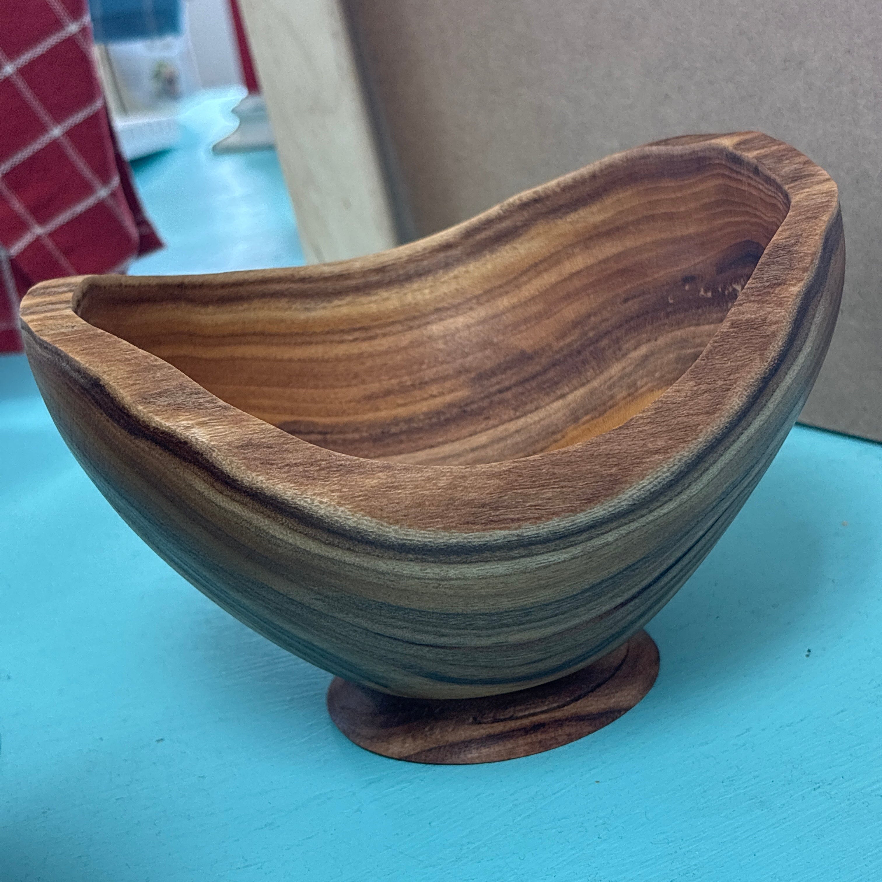 A handcrafted wooden bowl with a unique organic shape, featuring rich brown and tan wood grain patterns, resting on a light blue surface.