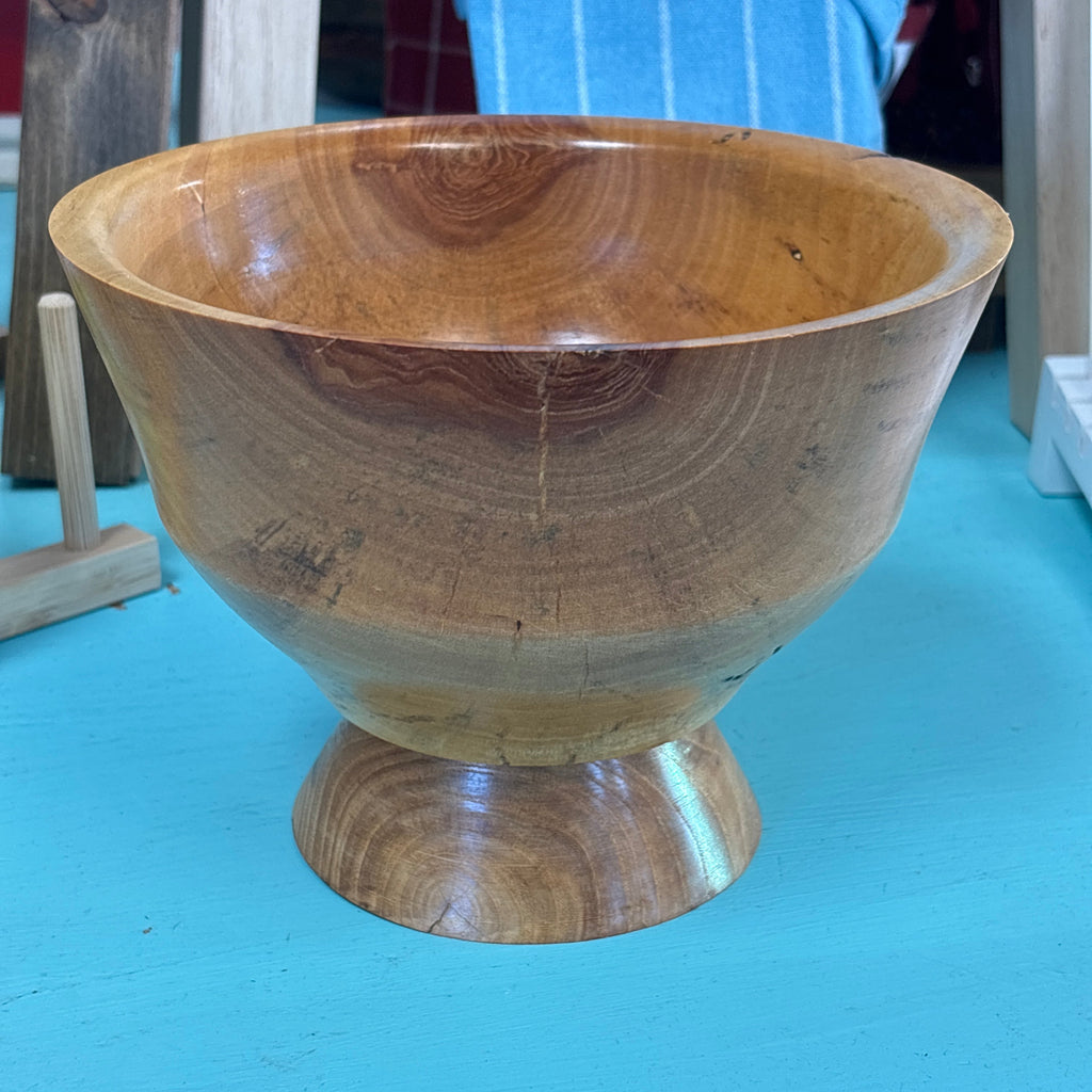 A handcrafted wooden pedestal bowl with a rich warm brown grain pattern, set against a bright turquoise background.