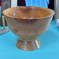 A handcrafted wooden pedestal bowl with a rich warm brown grain pattern, set against a bright turquoise background.