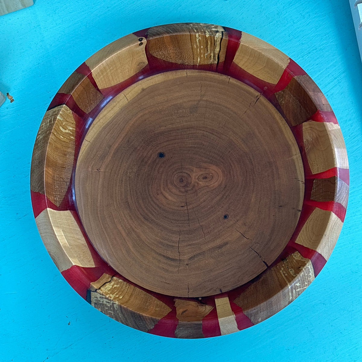 A round wooden bowl with a natural wood grain center and a rim made of alternating light and dark wood strips with red accents, shown from a top-down view on a bright blue surface.