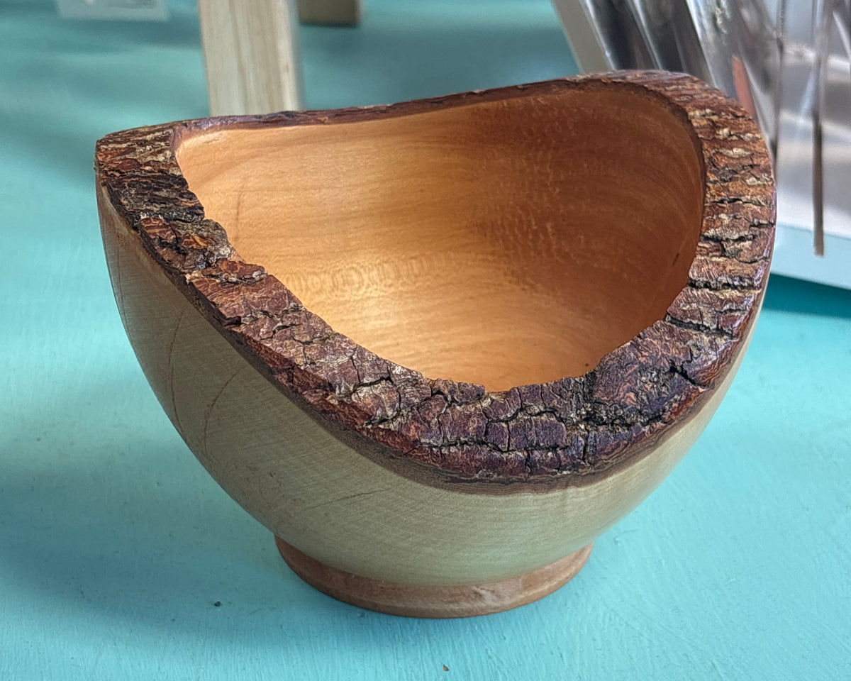 A handcrafted wooden bowl with a natural bark edge and a small footed base, shown on a turquoise tabletop with a blurred display stand in the background.