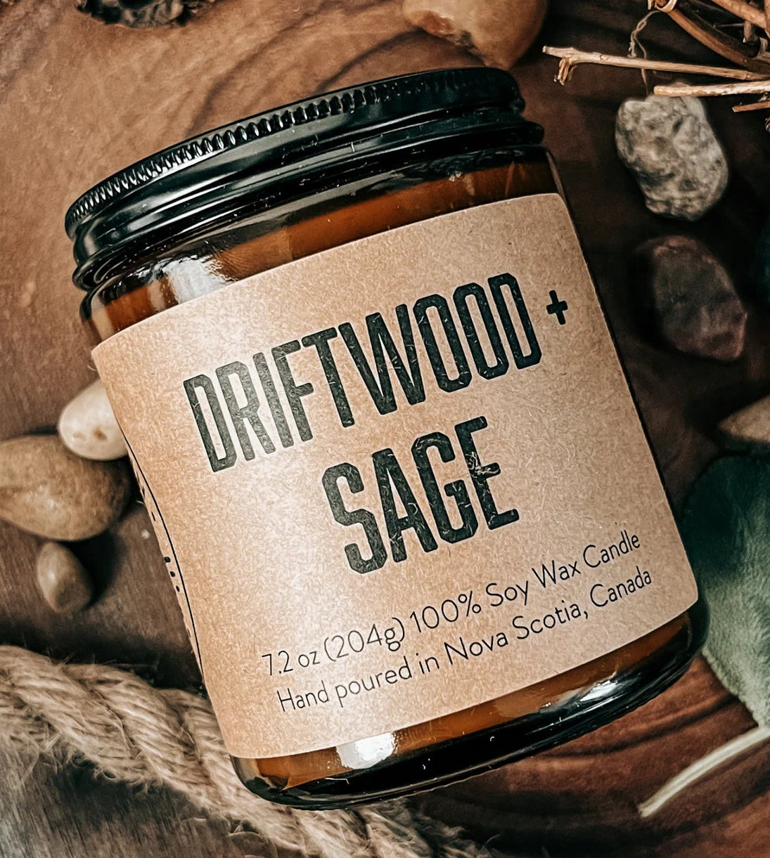 A jar candle labeled 'DRIFTWOOD + SAGE' sits on a rustic wooden tray surrounded by dried botanicals, green leaves, small stones, and rope. The candle jar has amber glass, a black lid, and a kraft-paper style label with black text.