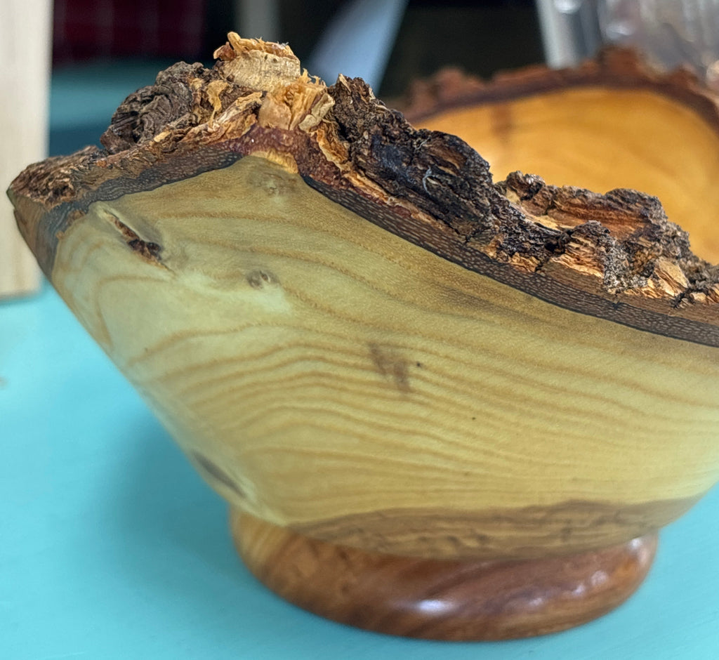 A handcrafted wooden bowl with a natural live edge and bark, displayed on a turquoise surface with a blurred background.