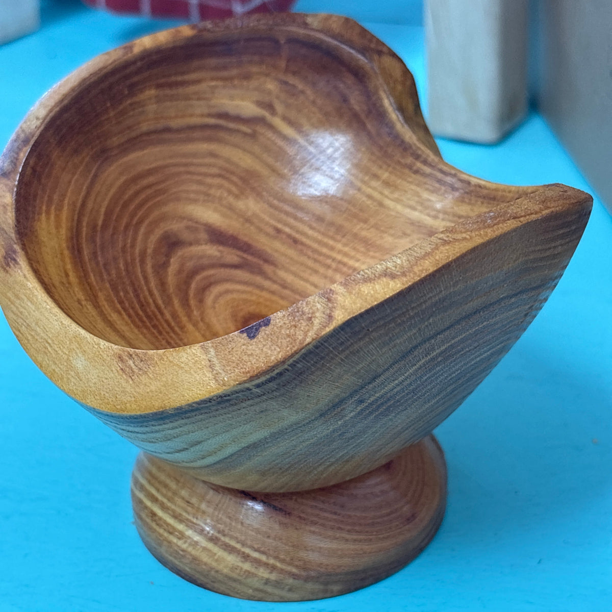 A handcrafted wooden bowl with a pedestal base, showcasing rich natural wood grain patterns in warm brown and golden tones, set against a bright blue background.