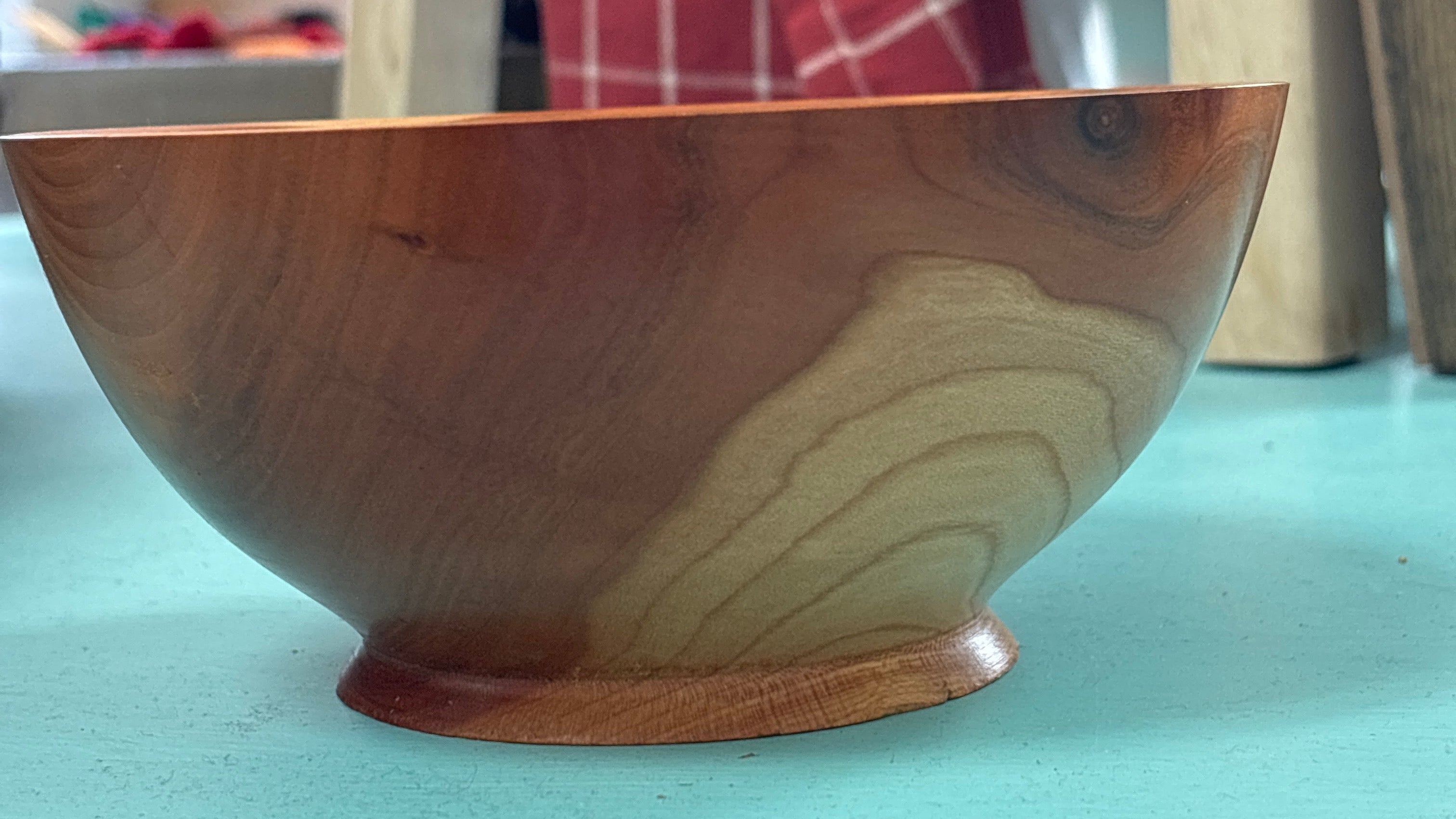 A handcrafted wooden bowl with a rich warm brown finish and natural wood grain patterns, sitting on a turquoise surface with a blurred background showing a wooden chair and a red checkered cloth.