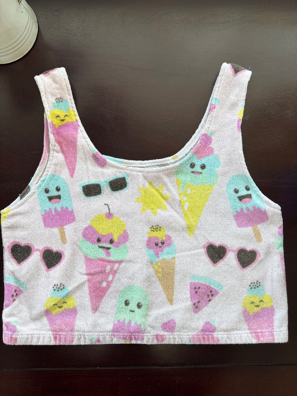 A white baby tank top  laid flat on a dark brown wooden surface, featuring a colorful pattern of cute ice cream cones, popsicles, watermelon slices, and hearts.