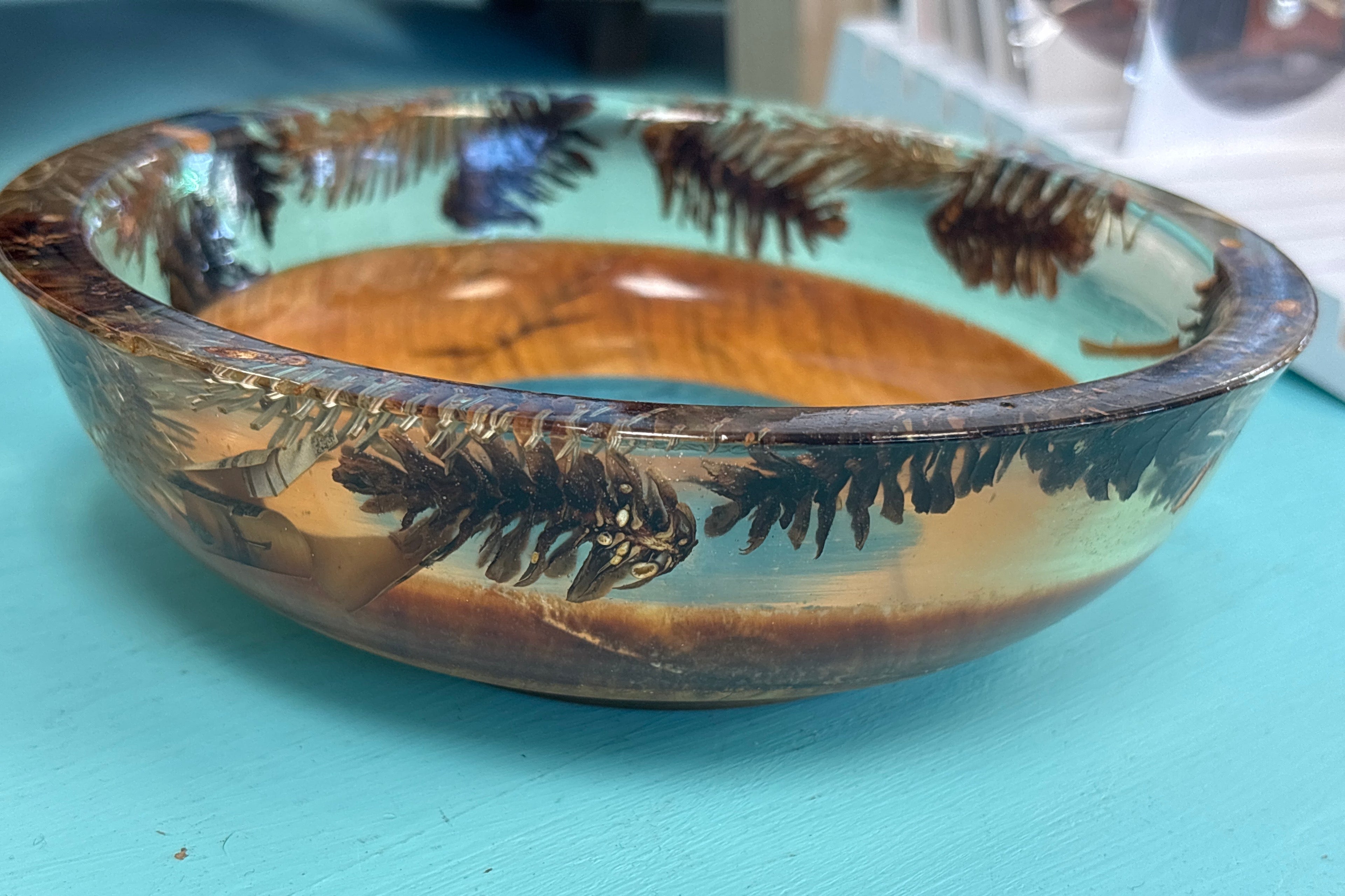 A handmade wooden bowl with a clear resin overlay containing dark brown pine needles, set against a turquoise painted wooden surface.