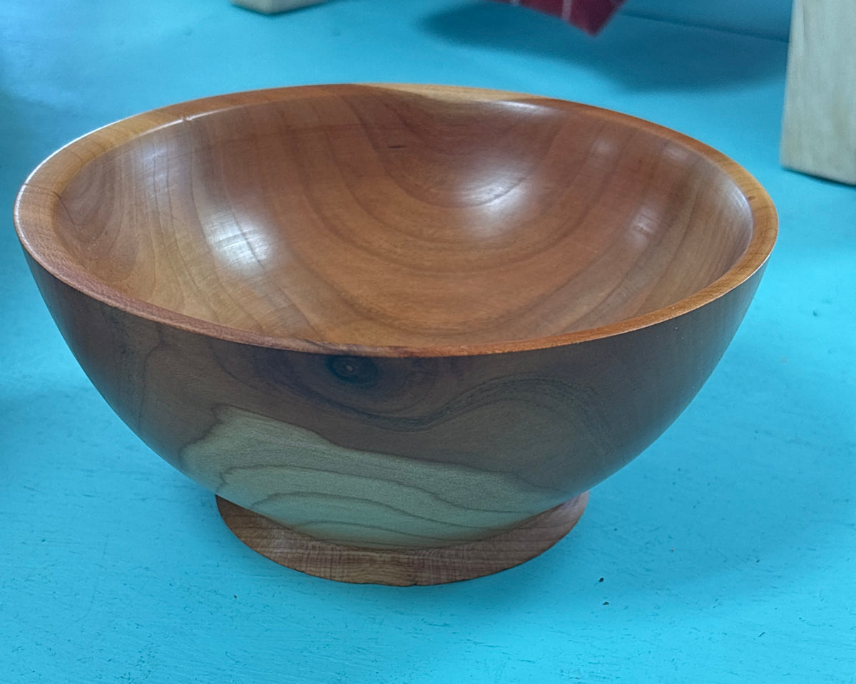 A handcrafted wooden bowl with a footed base, showcasing natural wood grain patterns in warm brown and reddish tones, set against a bright turquoise background.
