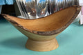 A rustic handcrafted wooden bowl with a live edge and a matching wooden pedestal base, displayed on a turquoise counter in a shop setting.