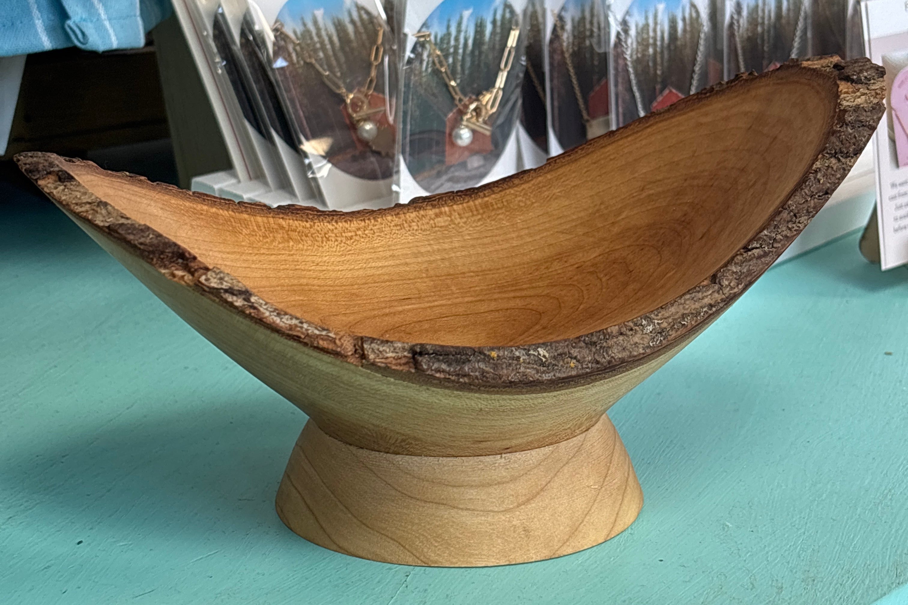 A rustic handcrafted wooden bowl with a live edge and a matching wooden pedestal base, displayed on a turquoise counter in a shop setting.