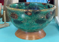 A round ceramic bowl with a glossy teal and green glaze, decorated with brown pinecones and gold glitter accents, resting on a wooden pedestal base.