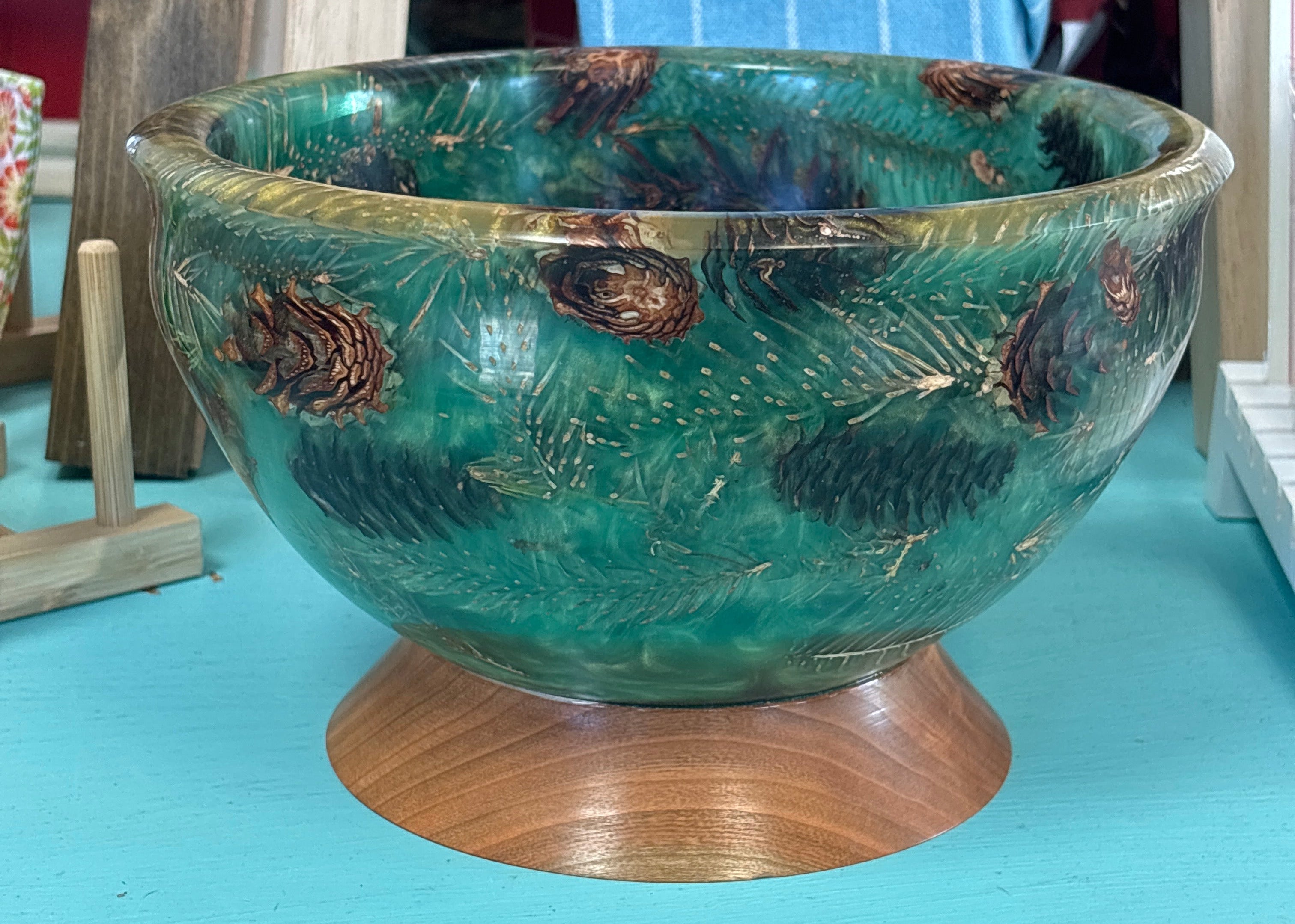 A round ceramic bowl with a glossy teal and green glaze, decorated with brown pinecones and gold glitter accents, resting on a wooden pedestal base.