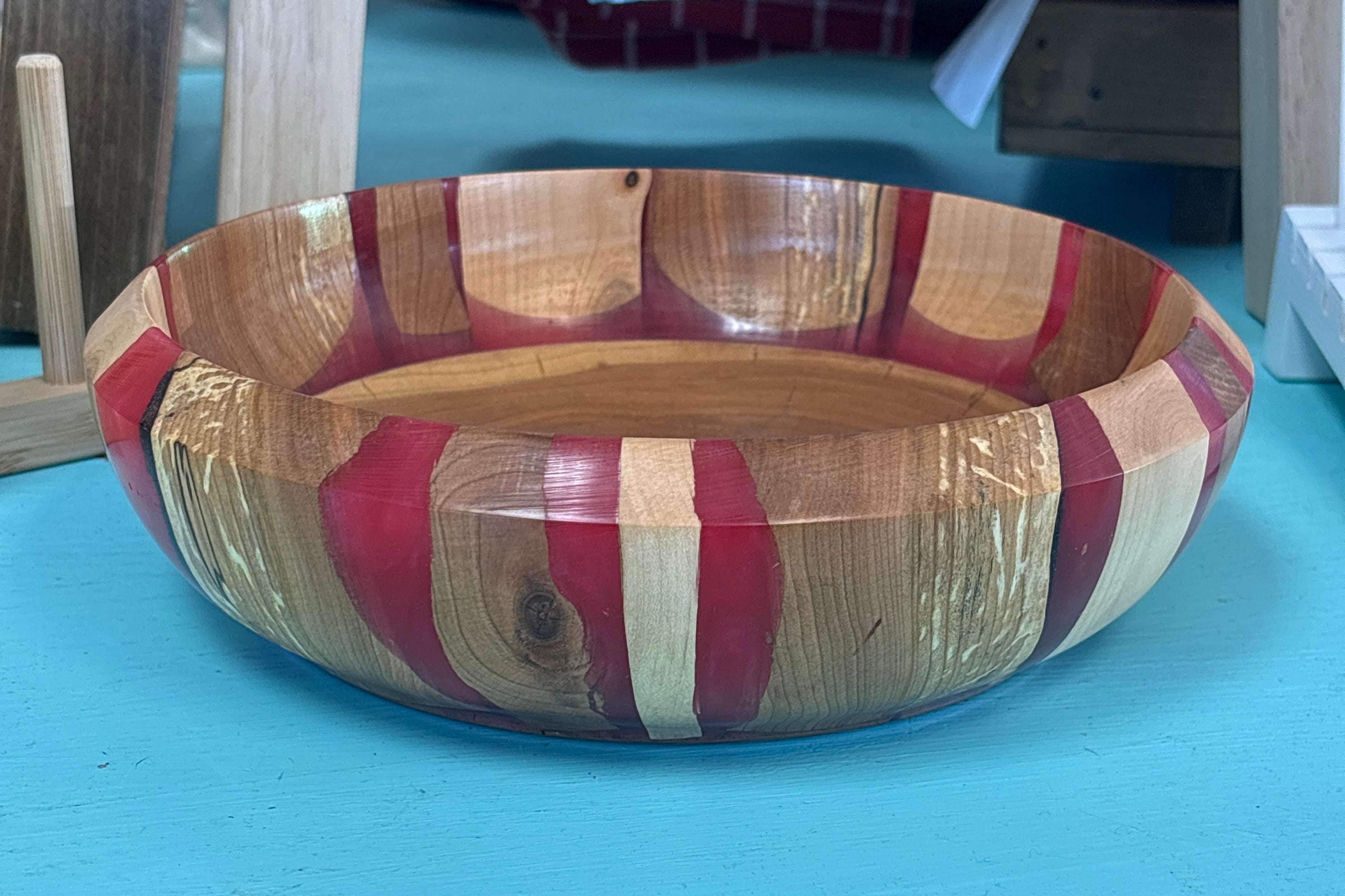 A handmade wooden bowl with alternating red, beige, and brown striped wood patterns, sitting on a bright turquoise painted table.