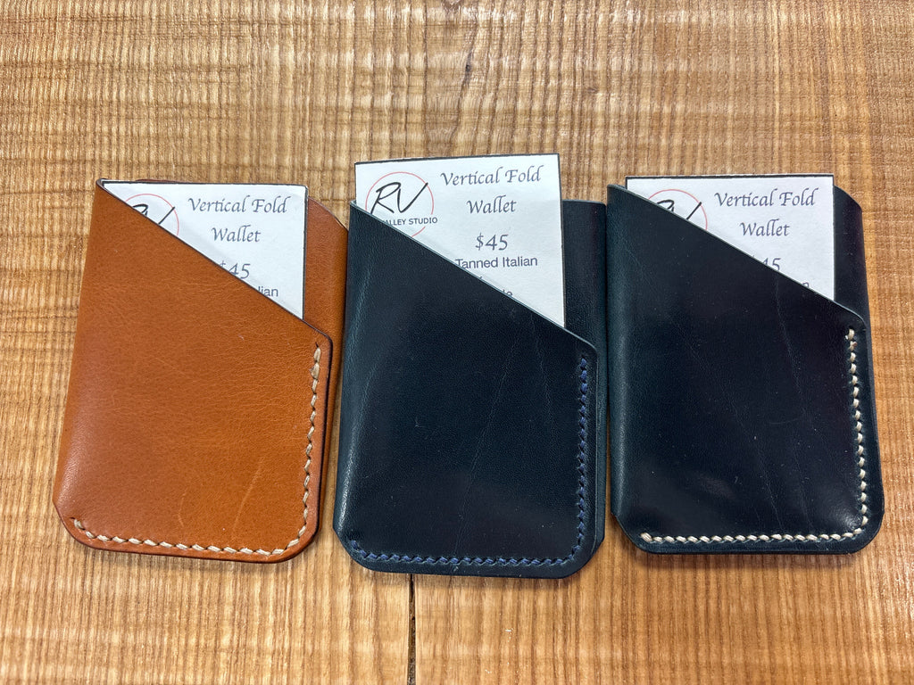 Three vertical fold wallets in brown, black, and black with white stitching on a wooden surface.