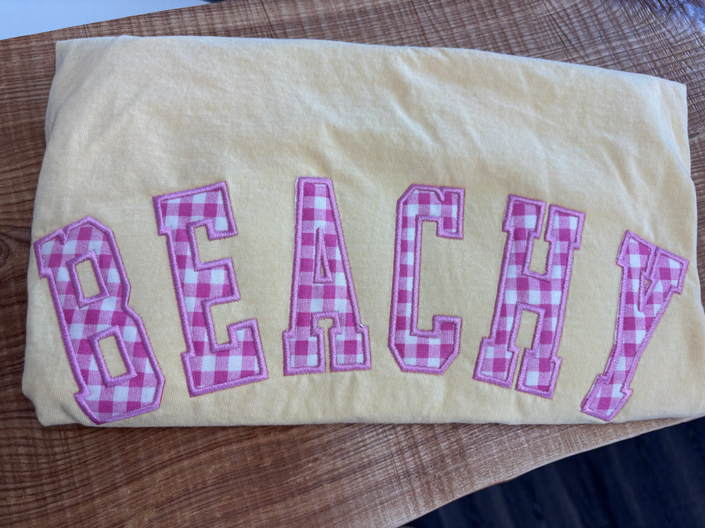 BUTTER COMFORT COLORS TSHIRT with 'BEACHY' in pink checkered letters on a wooden surface