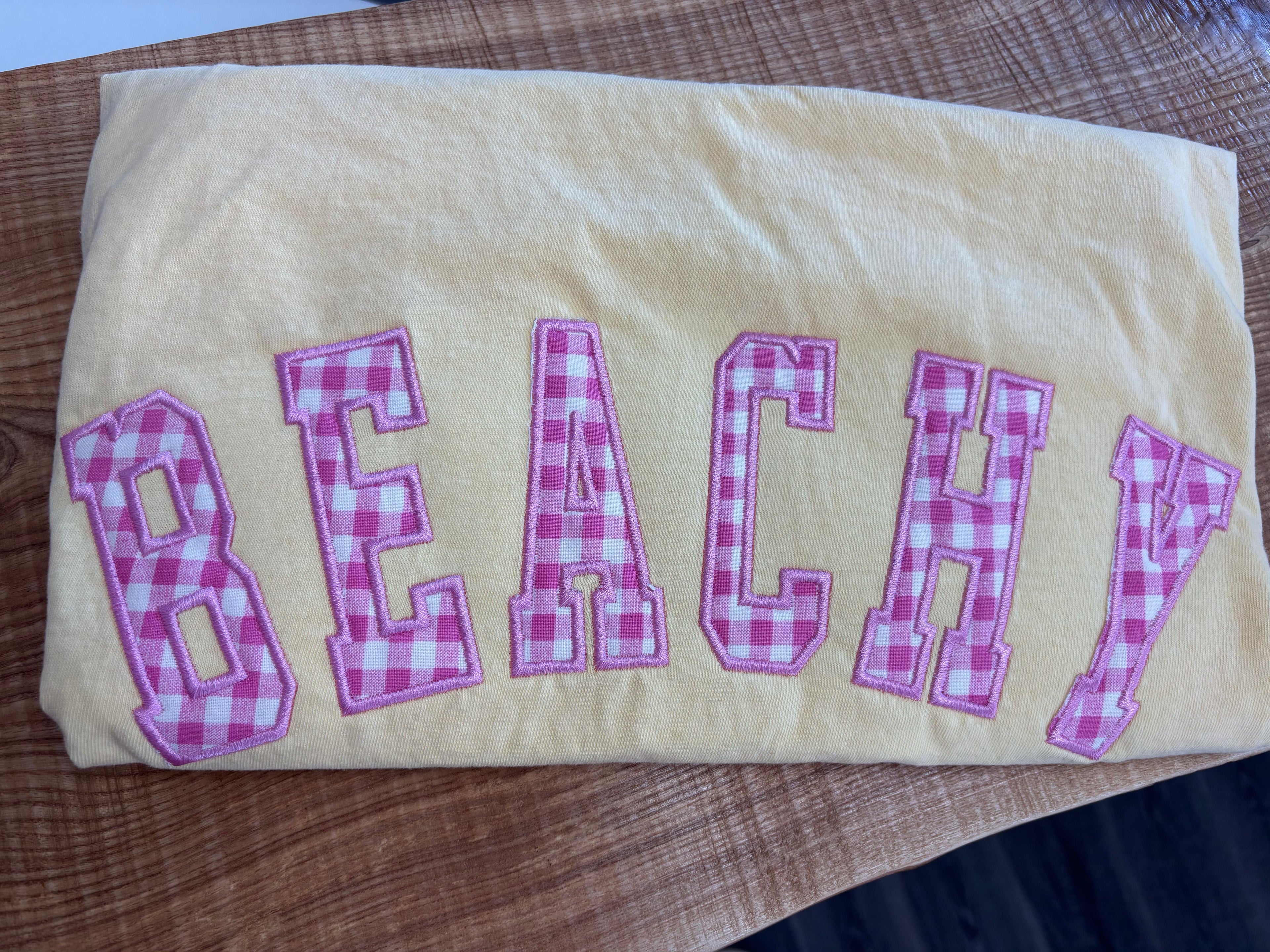 BUTTER COMFORT COLORS TSHIRT with 'BEACHY' in pink checkered letters on a wooden surface