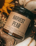 A jar candle labeled 'HARVEST PEAR' styled in a flat lay with a pear, sunflower, pinecones, cinnamon sticks, star anise, and dried leaves on a burlap surface.