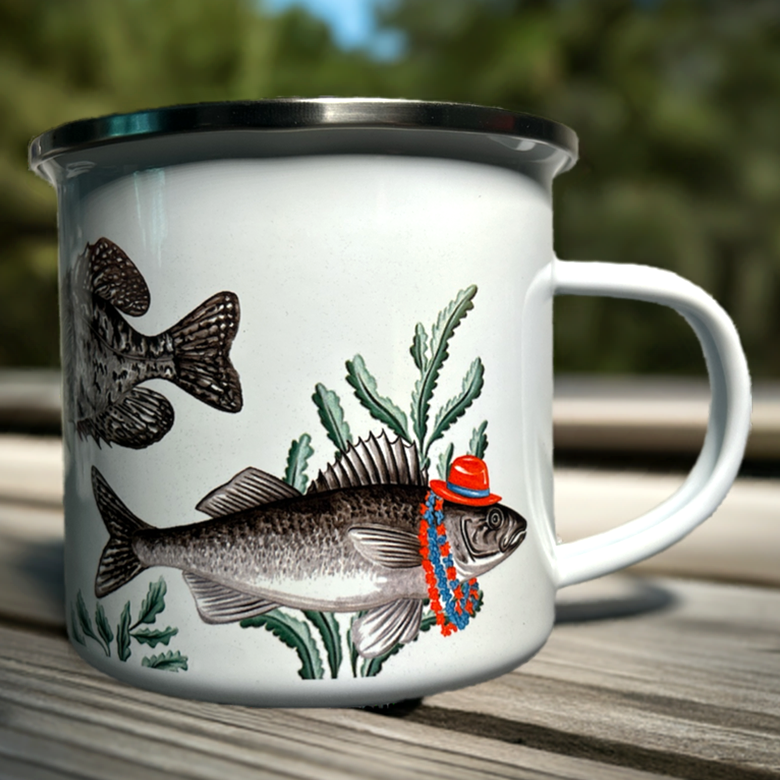White enamel mug with fish and nature-themed design on a wooden surface with blurred greenery in the background