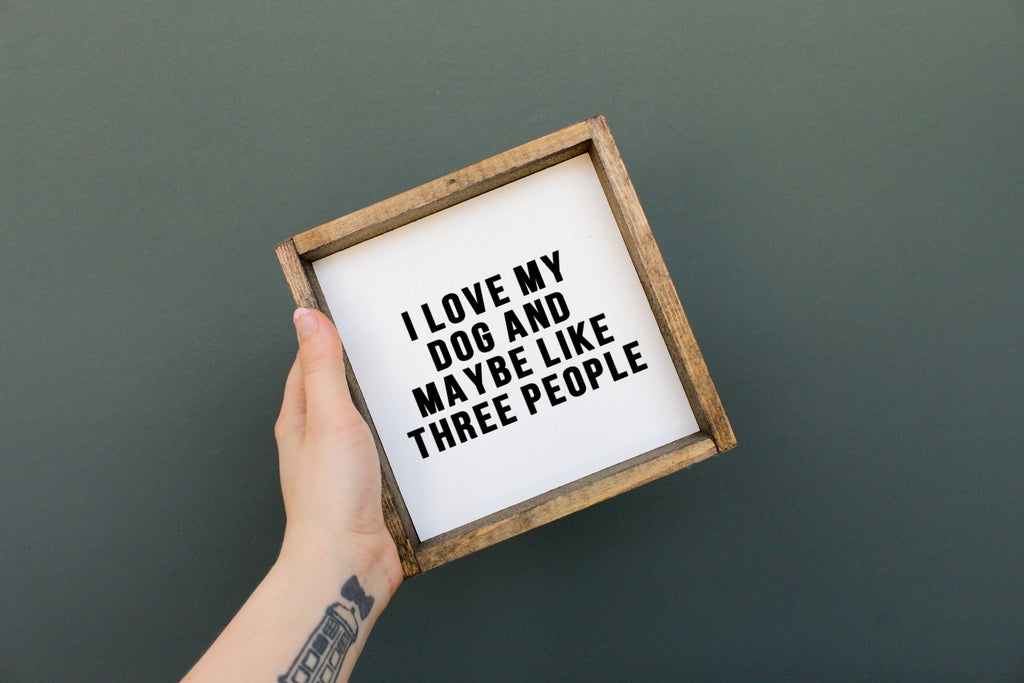 Hand holding a framed sign with text 'I love my dog and maybe like three people' against a dark background