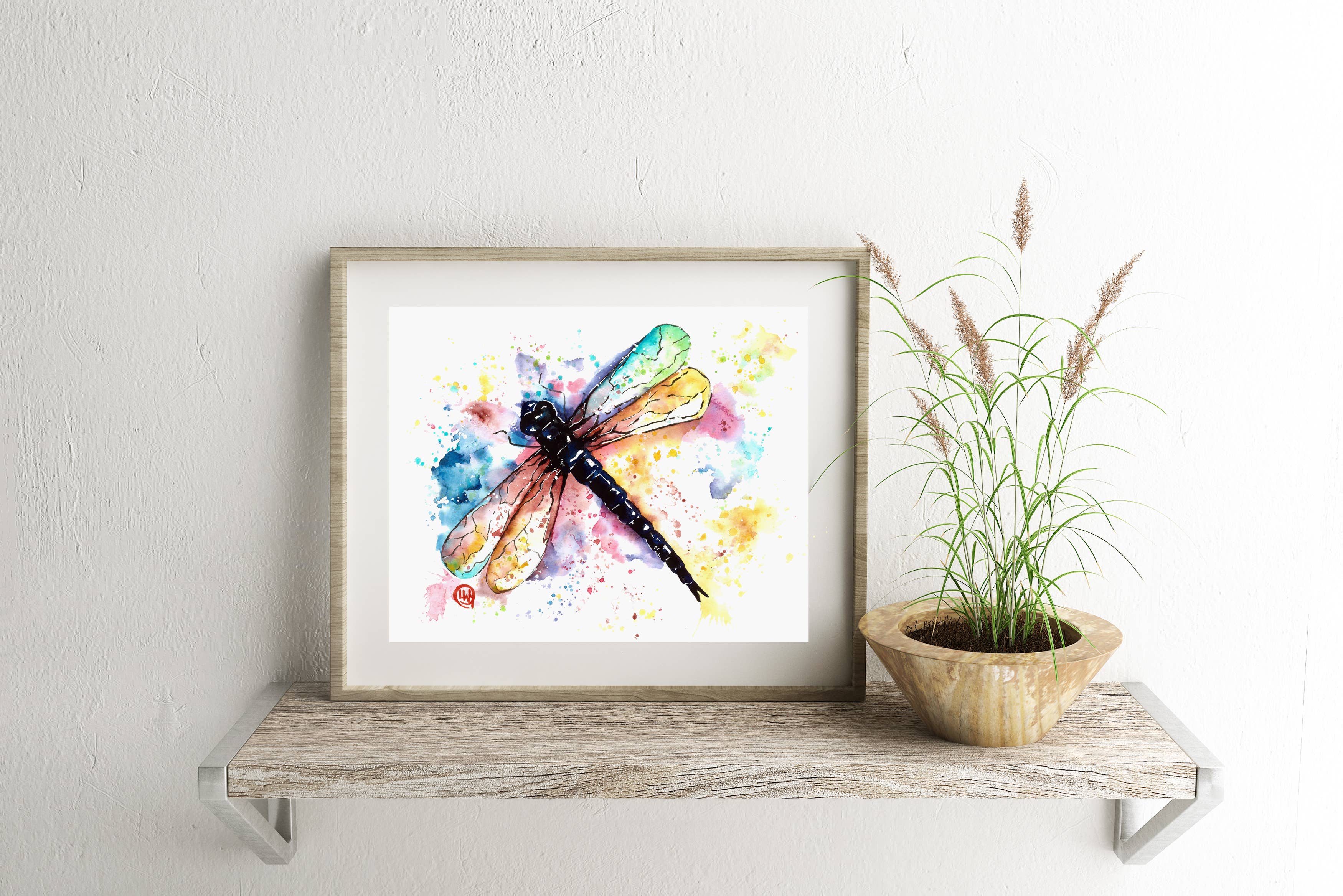 Framed artwork of a colorful dragonfly on a wooden shelf with a plant.