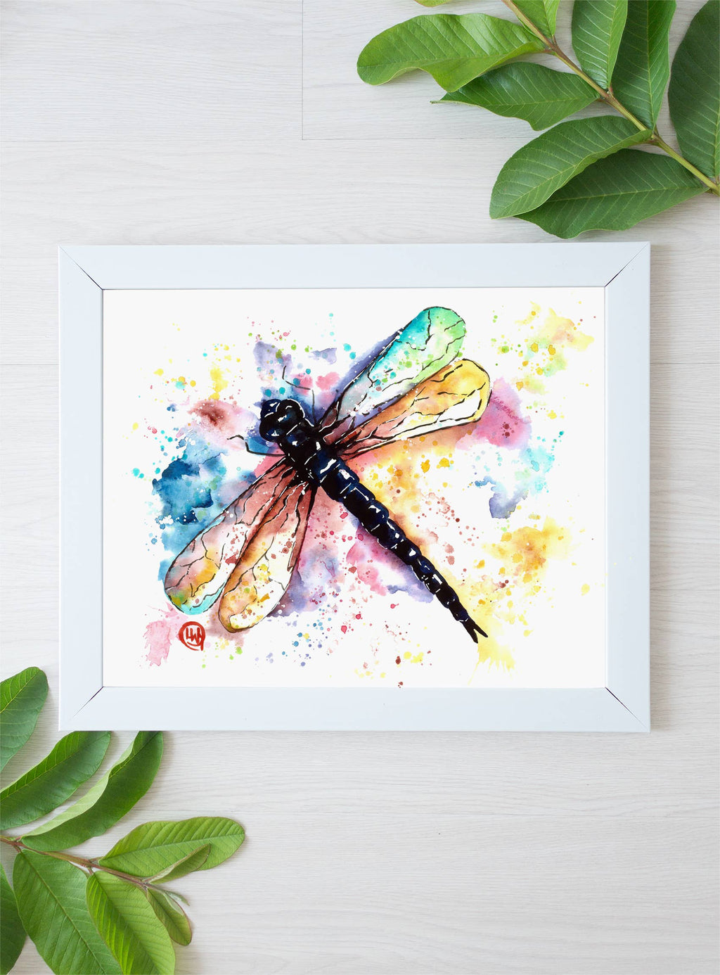 Framed watercolor painting of a dragonfly with colorful splashes on a white background.