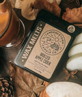 A styled flat-lay of a wax melt tin labeled 'Milled Apple Corp' by 'Timber Town Candle Co,' surrounded by dried apple slices, cinnamon sticks, star anise, pinecones, and a glass of amber drink on a burlap surface.