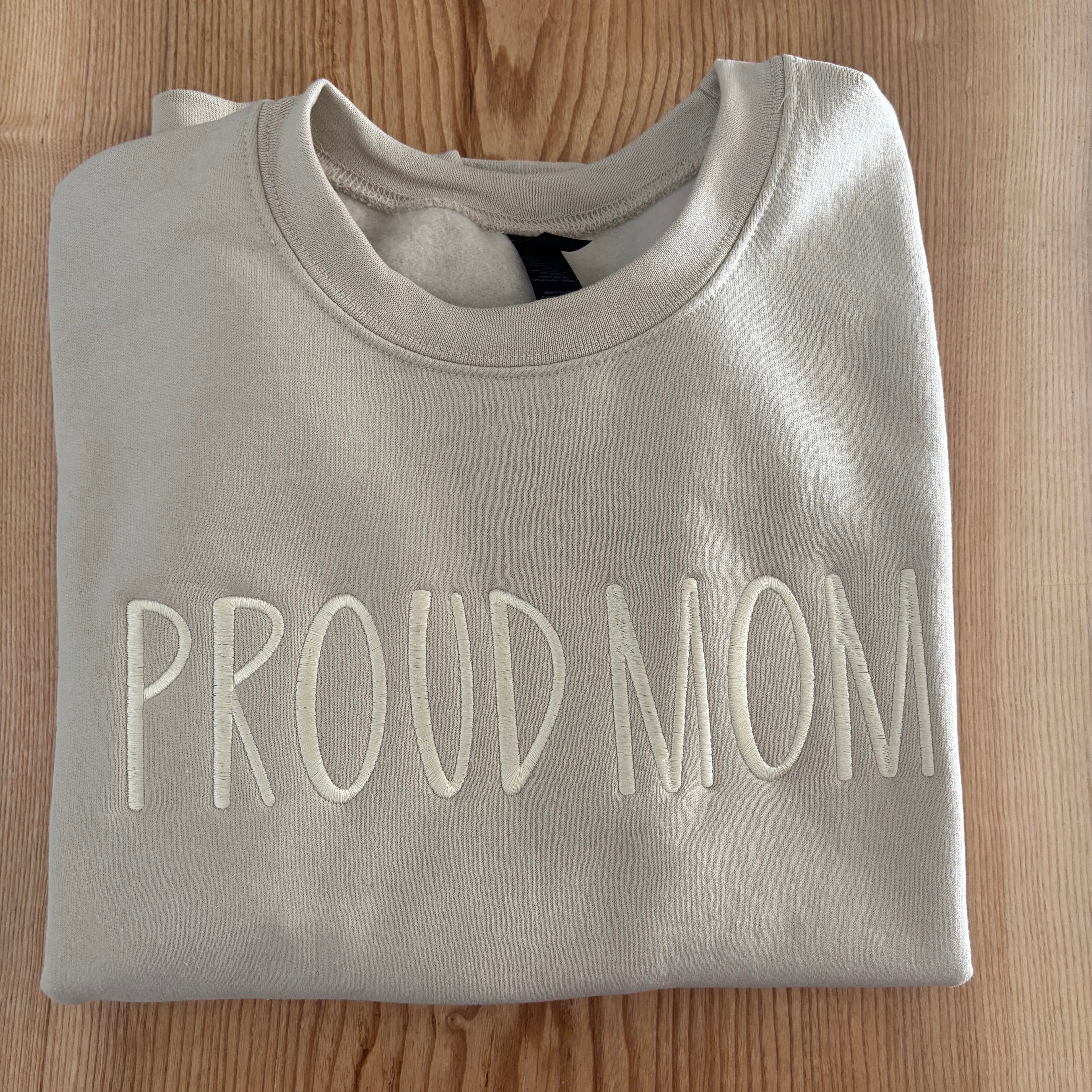 Light sand sweatshirt with 'PROUD MOM' text on a wooden surface
