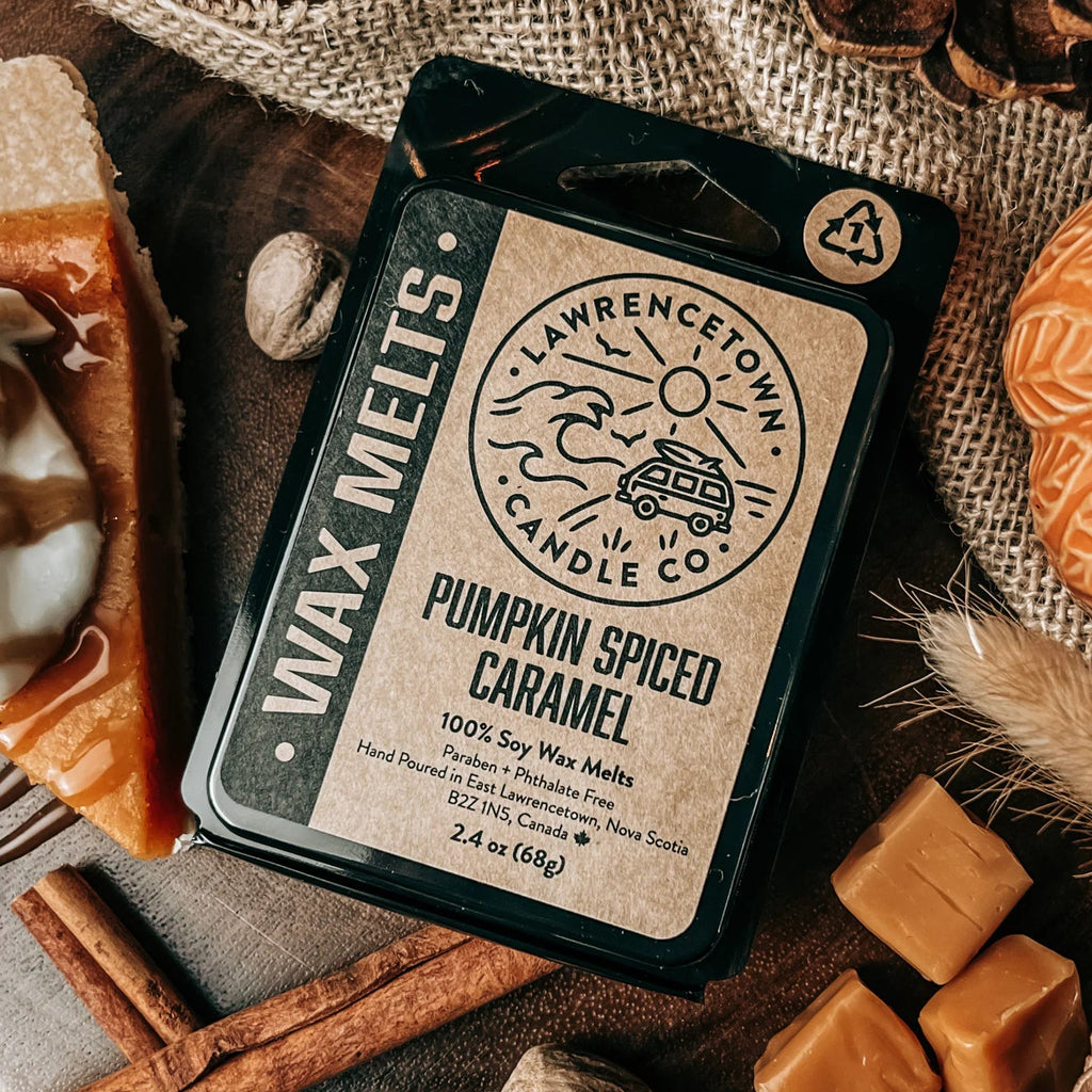 A package of Lawrence Town Candle Co. Pumpkin Spiced Caramel wax melts is displayed in a cozy autumn flat-lay setting with a lit candle, pinecones, cinnamon sticks, caramel squares, and a slice of pumpkin pie.