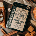 A package of Lawrence Town Candle Co. Pumpkin Spiced Caramel wax melts is displayed in a cozy autumn flat-lay setting with a lit candle, pinecones, cinnamon sticks, caramel squares, and a slice of pumpkin pie.